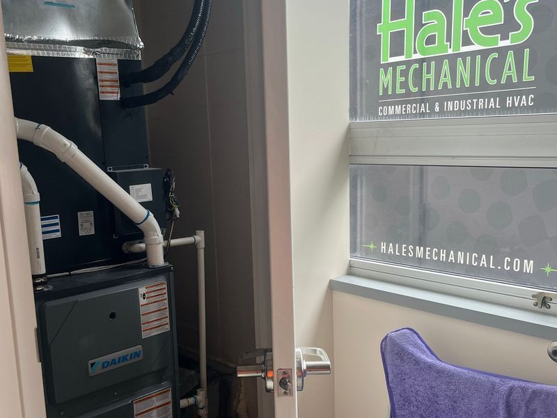 A Daikin HVAC unit in a small utility closet next to a glass window with a Hales Mechanical company advertisement.