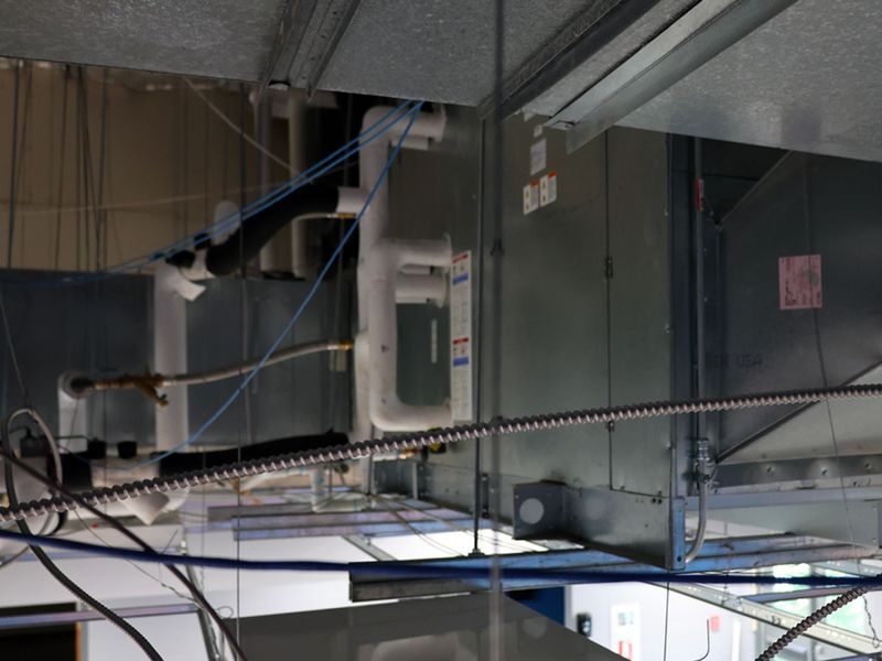 An industrial HVAC unit suspended from a ceiling with white plumbing, electrical wiring, and ductwork.