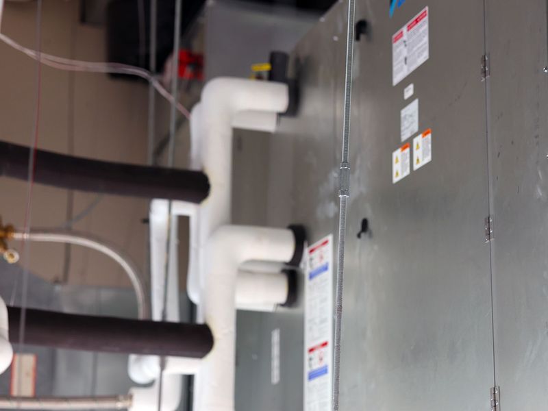 A close-up view of an indoor HVAC unit with insulated white pipes and metal casing featuring various warning labels.