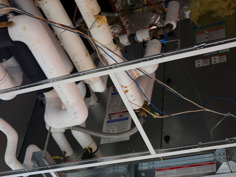 Ceiling-mounted HVAC unit with white insulated pipes and electrical wiring visible above a suspended ceiling grid.