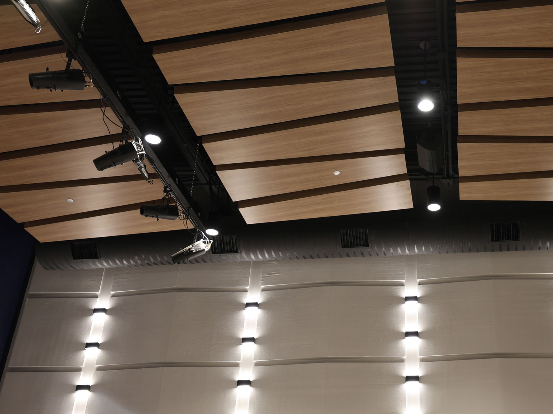 A low-angle view of a modern ceiling with wooden slats, black structural beams, stage lights, and wall-mounted sconces.