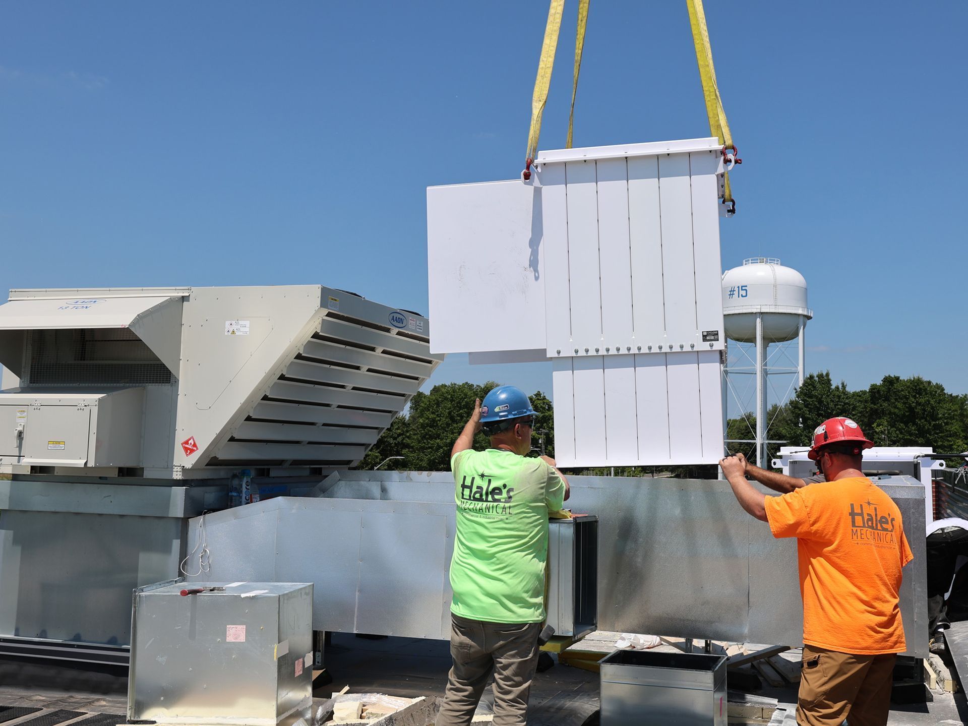 Two workers in high-visibility shirts guide a large, white HVAC unit being lowered by a crane onto a metal duct system.