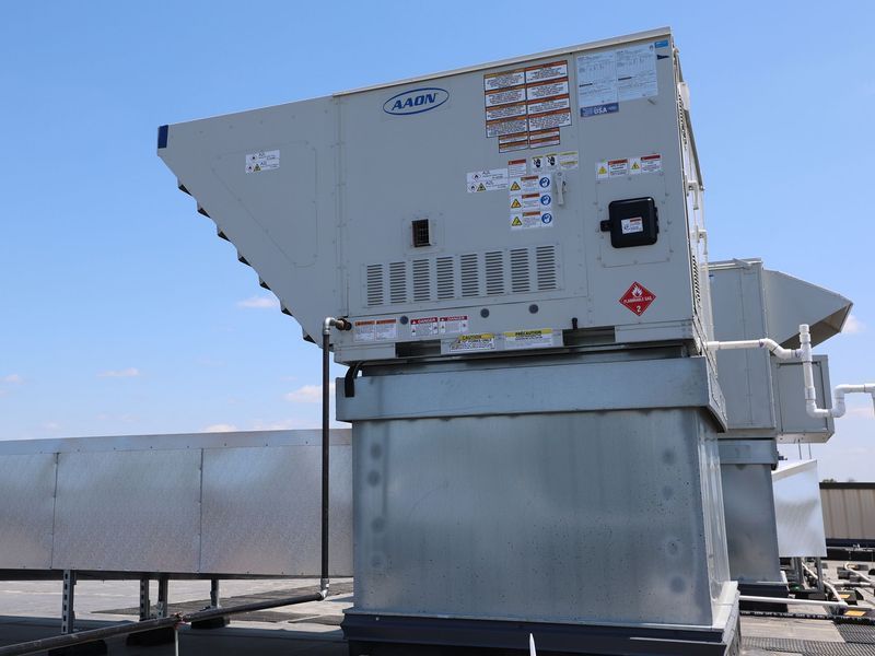 An industrial AAON rooftop HVAC unit and metal ductwork sit under a bright blue sky.