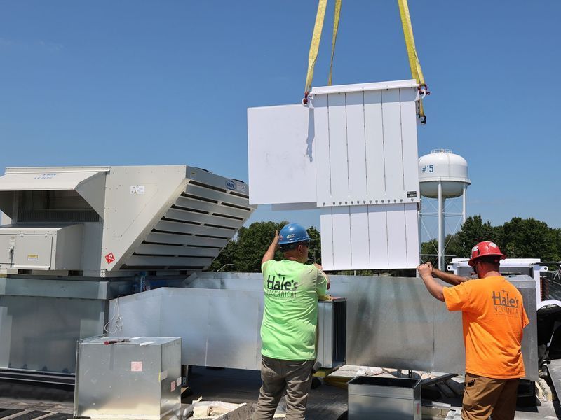 Two construction workers in high-visibility shirts guide a large, white HVAC unit being lowered by a crane onto a roof.