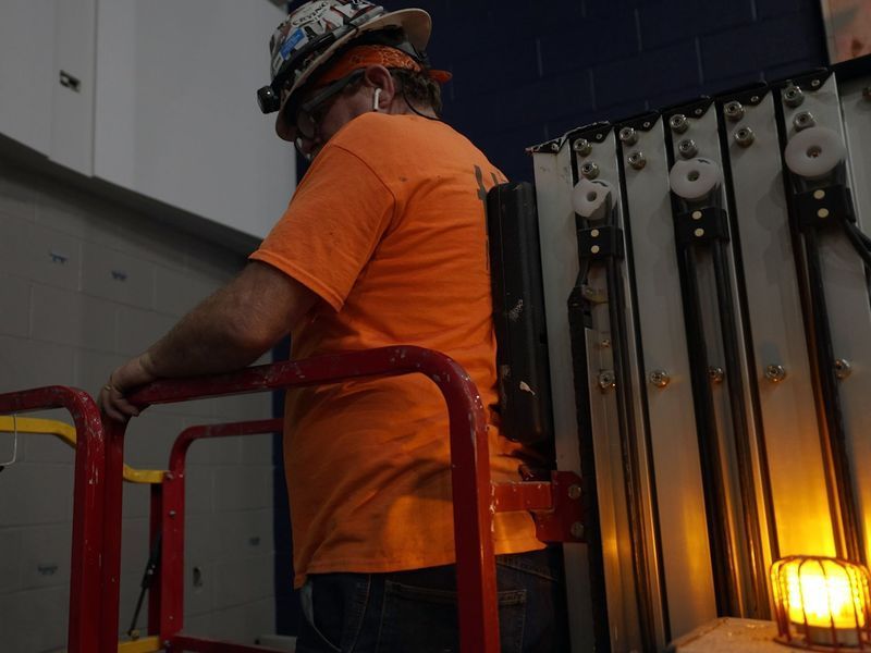 A person in a hard hat and orange shirt operates an aerial lift, leaning against the machinery inside a building.