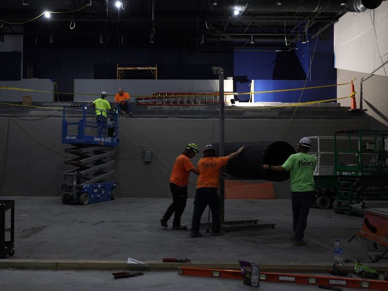 Construction workers in high-visibility vests work on a large pipe inside an unfinished, indoor commercial space.
