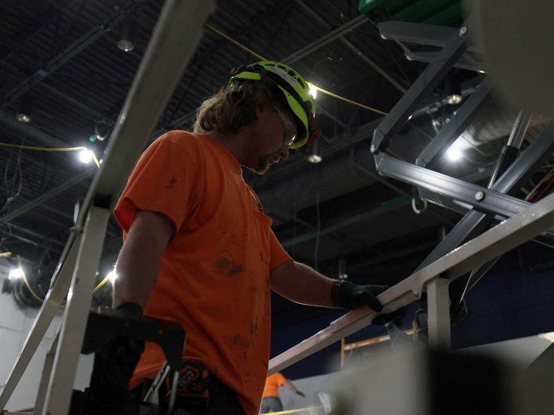 A person in a bright orange shirt and a yellow hard hat working on metal structures inside a construction site.
