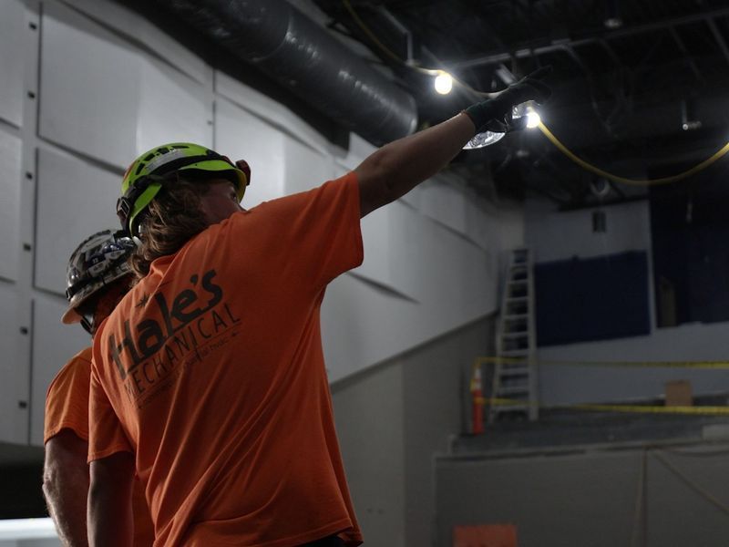 Two workers in orange shirts and hard hats point upward at exposed ceiling infrastructure in an indoor work site.