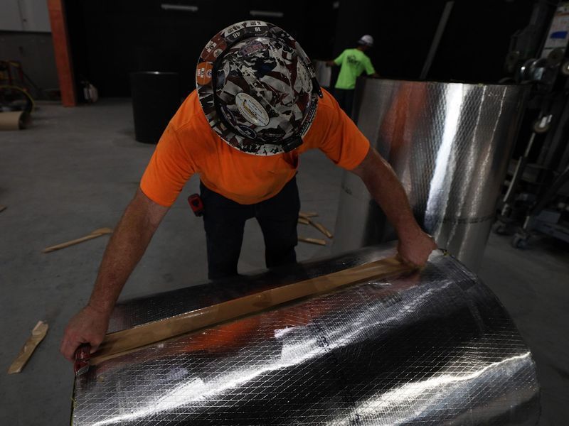 A person in an orange shirt and patterned hard hat uses a wooden straightedge to mark a roll of reflective insulation.