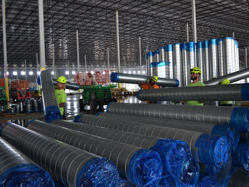 Workers in a warehouse carry large metal pipes near stacks of wrapped industrial conduits.