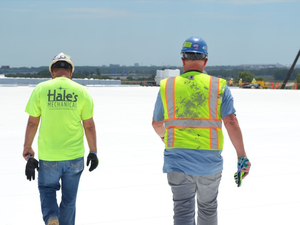 Two workers in hard hats and safety gear walk across a white flat rooftop on a sunny day.