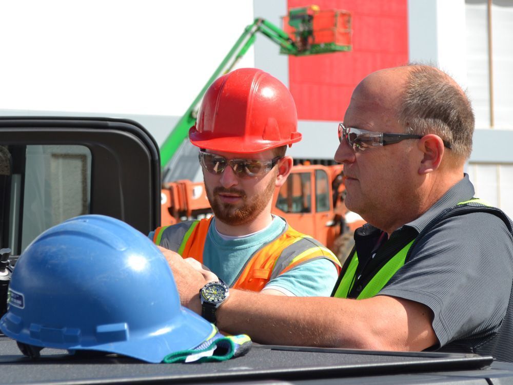 Two people wearing safety vests and glasses discuss work near a construction site; a blue hard hat sits in the foreground.