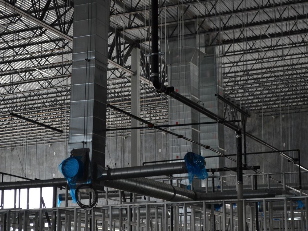 Industrial building interior showing vertical and horizontal metallic air ducts and dark pipes suspended from a truss roof.