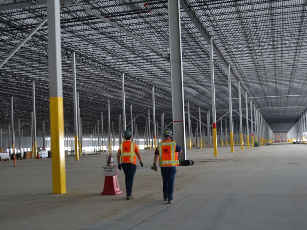 Two construction workers in high-visibility vests walk through a large, empty warehouse with high steel ceilings.