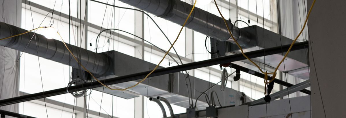 Exposed ceiling infrastructure featuring metal ventilation ducts, electrical conduit, and wires against a glass window wall.