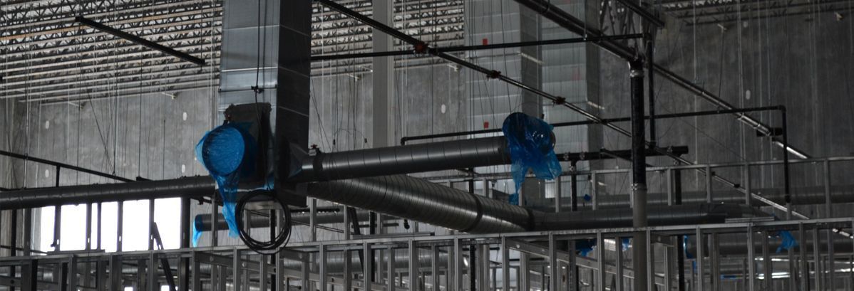 Metal ventilation ducts with blue protective plastic covers hanging from a ceiling frame in an unfinished building.