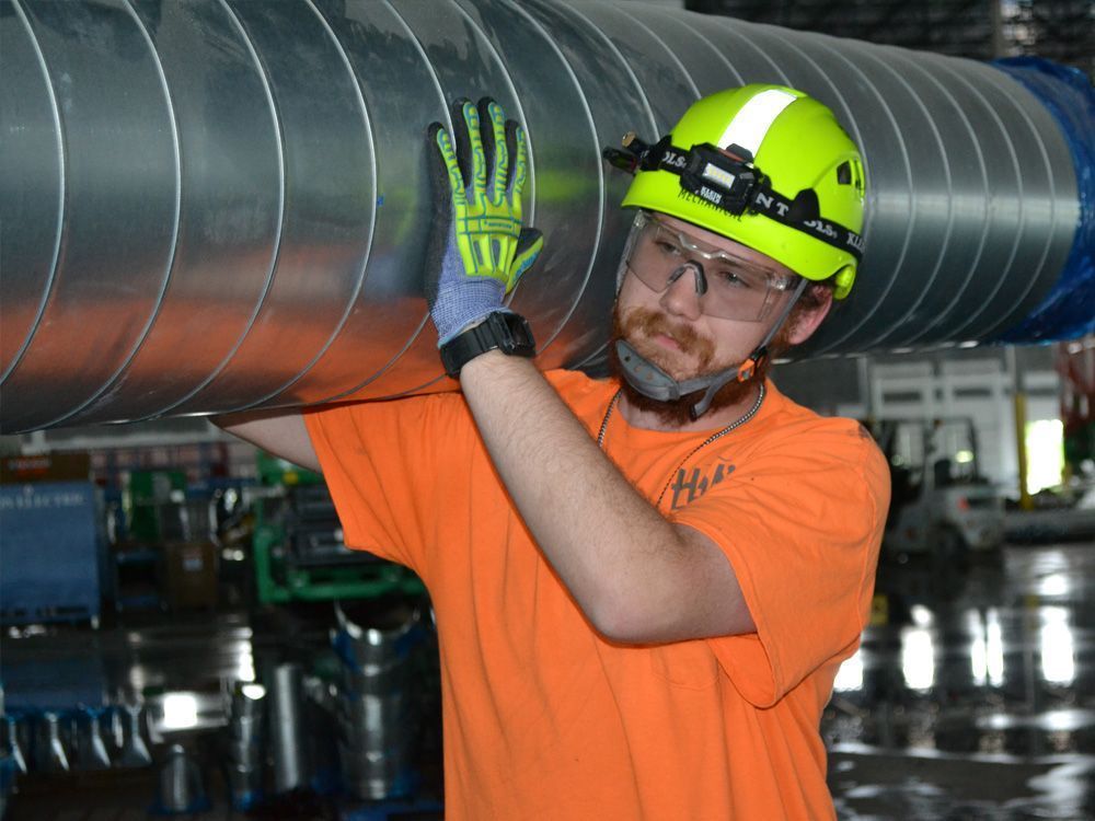 A worker wearing a lime yellow hard hat, safety glasses, and gloves carries a large, silver metal duct on their shoulder.