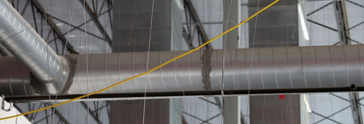 Metal HVAC ductwork suspended from the ceiling of a large industrial building with a yellow guide wire running across.