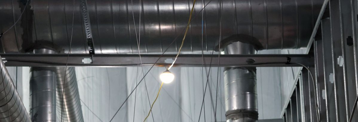 A low-angle view of industrial metal ductwork and a structural beam with a single, glowing pendant light hanging below.