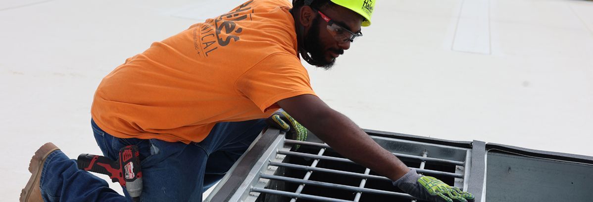 A construction worker in an orange shirt and hard hat kneels on a white roof, installing a metal vent cover.