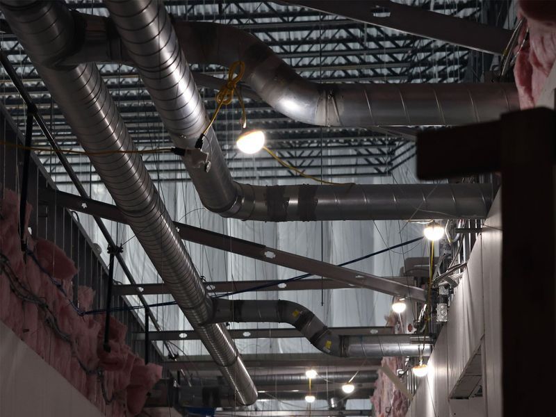 Metal ventilation ducts and lighting fixtures hanging from an open ceiling in an industrial building space.