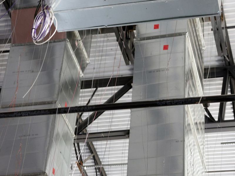 Two large, silver, rectangular HVAC ducts suspended from a white metal ceiling with exposed steel beams and wiring.