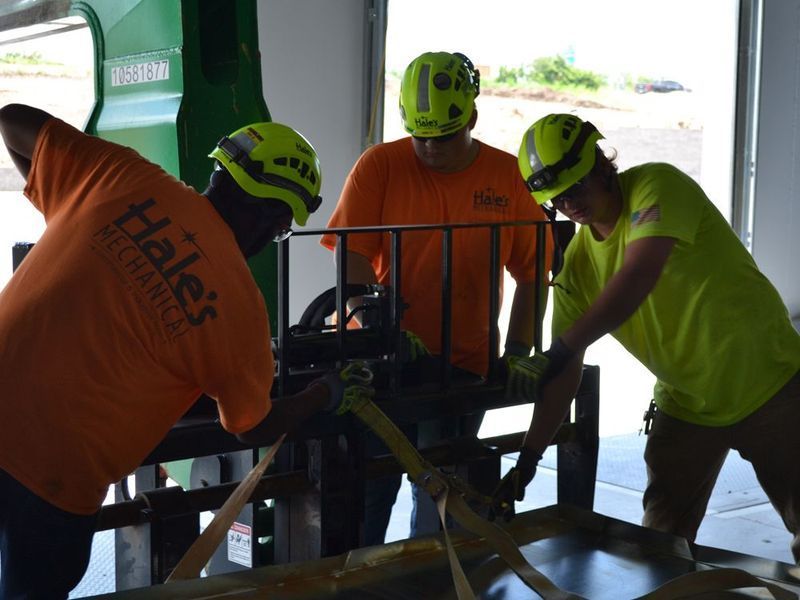Three people in safety helmets and bright shirts work together to secure straps on equipment in an industrial setting.