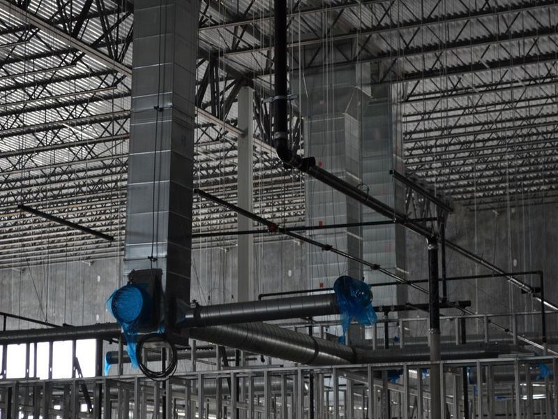 Construction site view of vertical metal air ducts and exposed ceiling joists with blue protective film on piping ends.