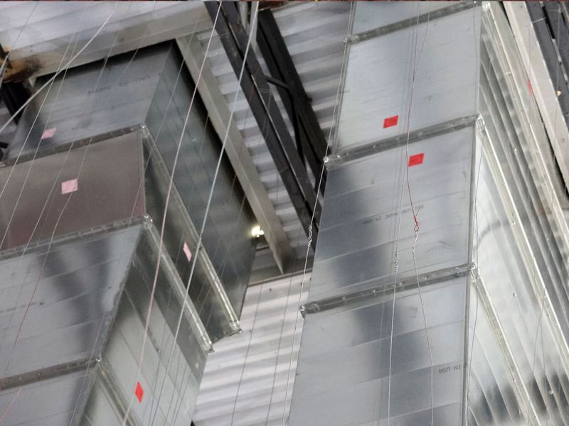 Rectangular metal HVAC ducts suspended from a ribbed ceiling by thin wires in a commercial or industrial space.