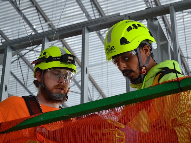 Two construction workers in high-visibility gear and hard hats consult while working on a project in an indoor structure.