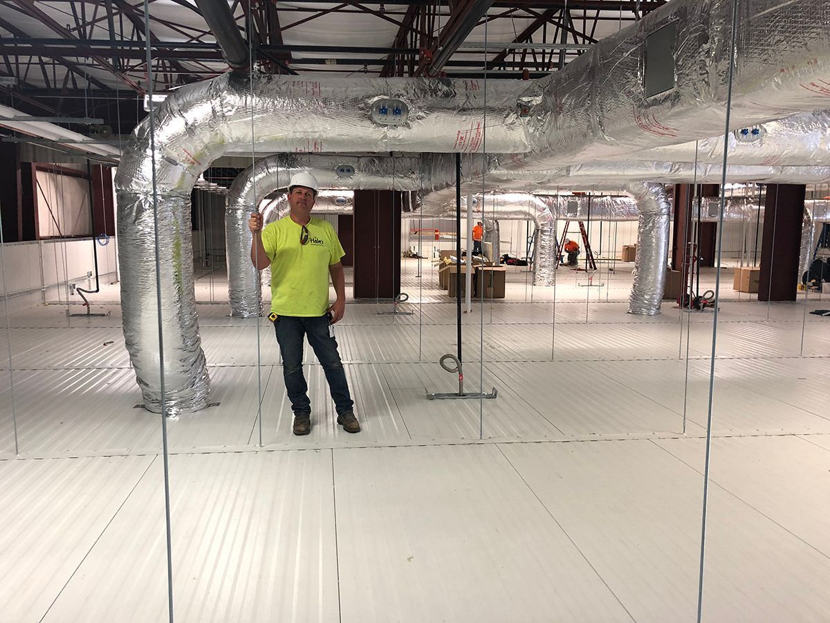 A construction worker in a high-visibility yellow shirt stands in a large interior space under exposed silver ductwork.