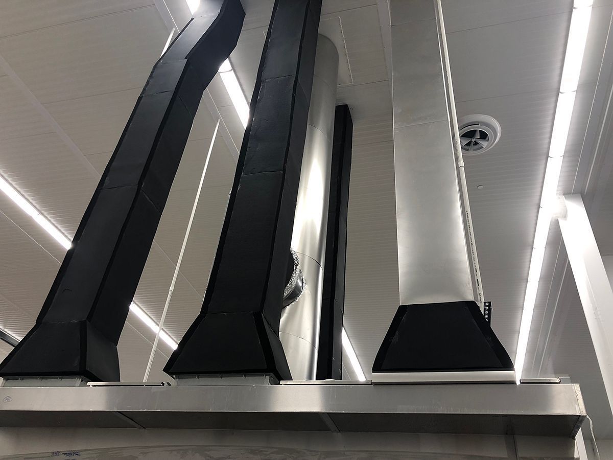 Three industrial ventilation ducts, two black and one silver, extending upward from a metal vent hood toward a ceiling.