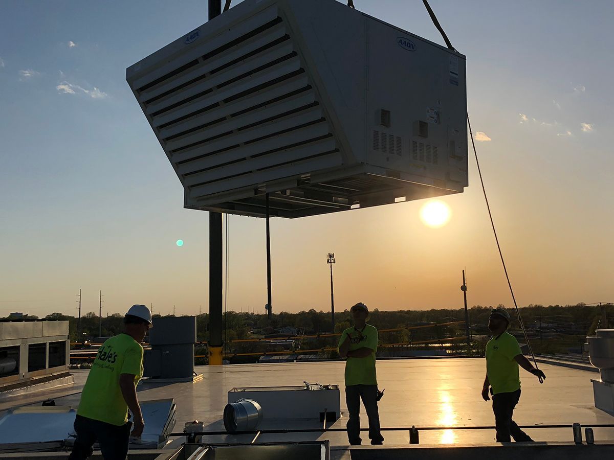 Construction workers on a rooftop watch as a crane lifts a large industrial HVAC unit at sunset.