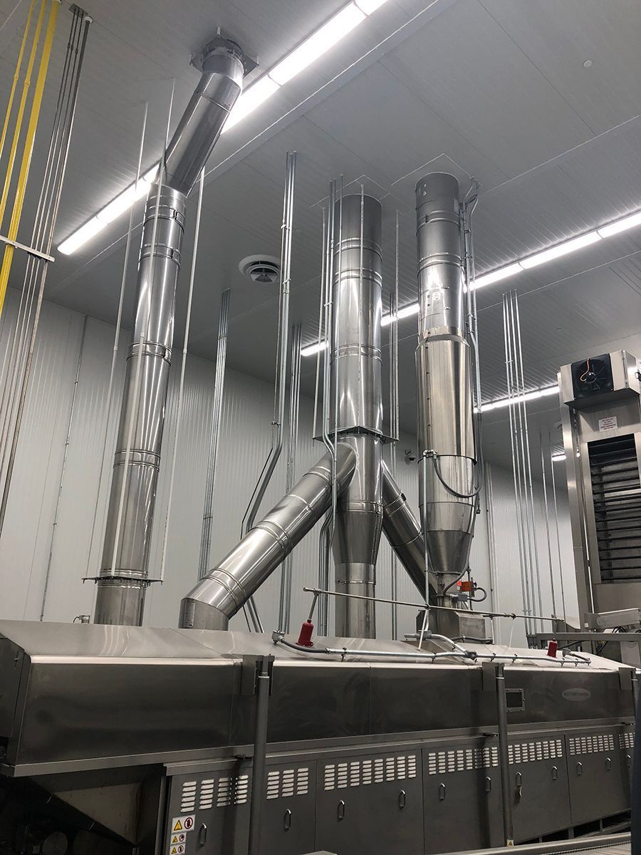 Industrial stainless steel exhaust ductwork and machinery inside a facility with white panel walls.