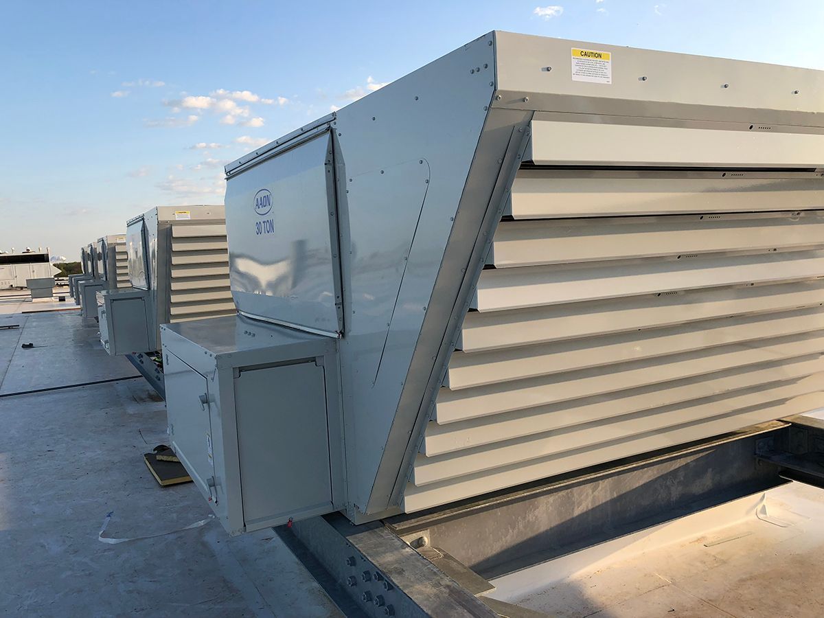 A row of grey industrial HVAC rooftop units installed on a flat, light-colored roof under a clear blue sky.