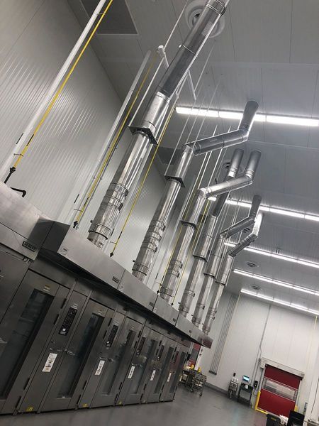 A row of industrial ovens in a large commercial kitchen, with multiple metal exhaust vents extending to the ceiling.