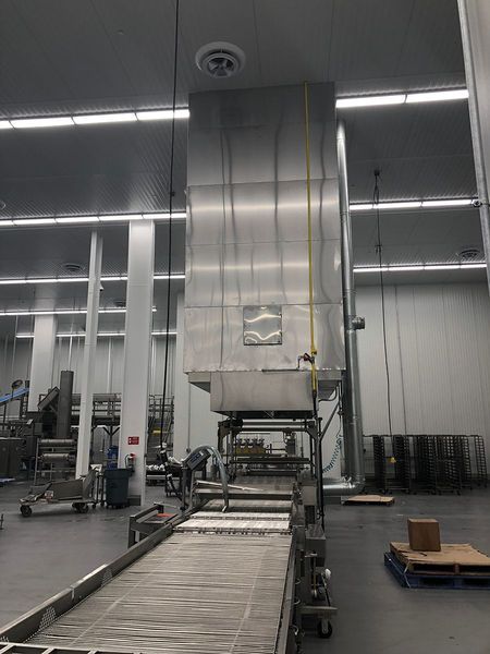 A large, stainless steel industrial vent hood hangs above a conveyor belt in a brightly lit, clean factory environment.