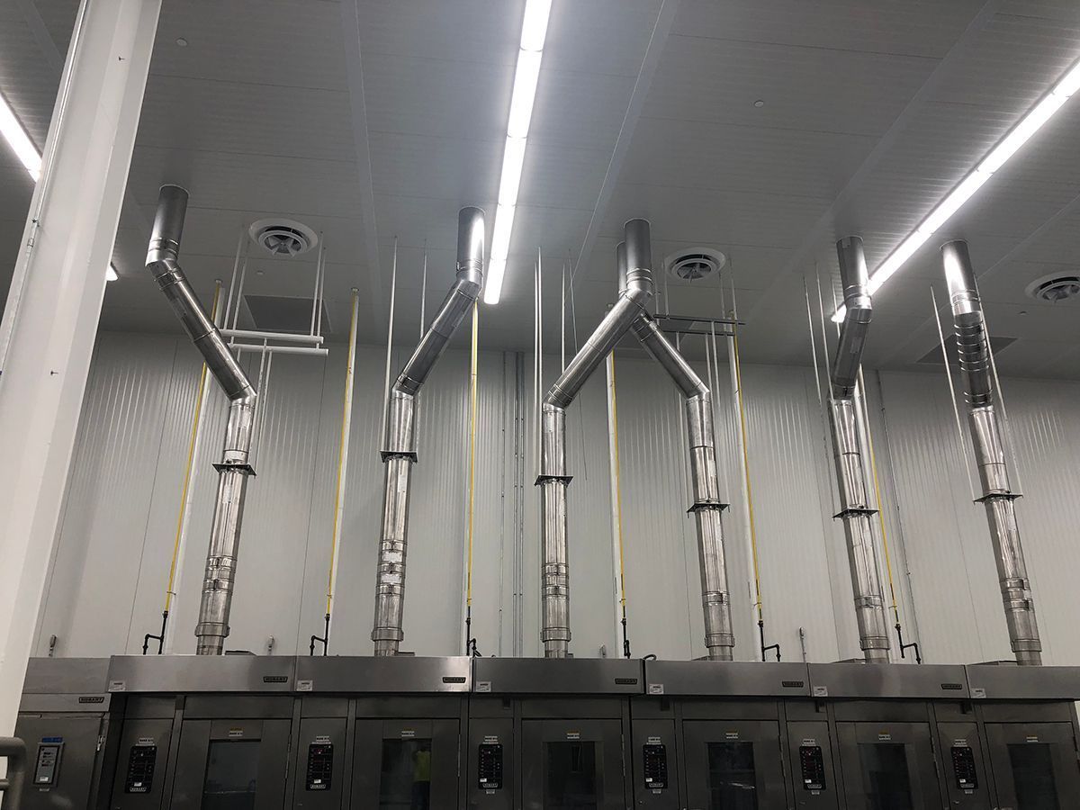 Stainless steel ventilation ducts and Y-shaped exhaust pipes connected to commercial kitchen ovens in a clean, bright room.