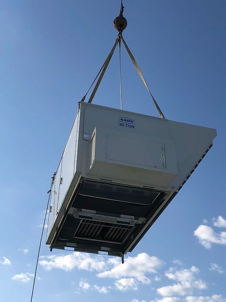 A large, industrial HVAC unit suspended in the air by a crane against a clear blue sky.