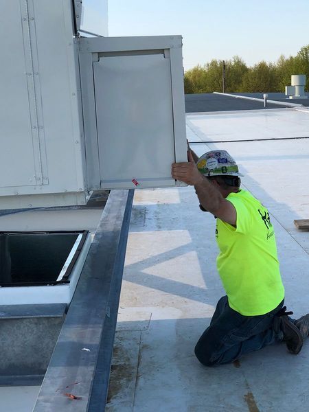 A worker in a bright yellow shirt and hard hat kneeling on a rooftop, adjusting a white metal ventilation panel.