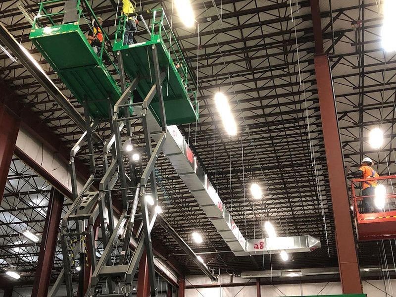 Workers on green and orange scissor lifts install overhead ductwork in a large industrial warehouse.