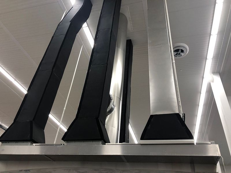 Low-angle shot of black and silver ventilation ducts extending from a metal hood against a brightly lit ceiling.