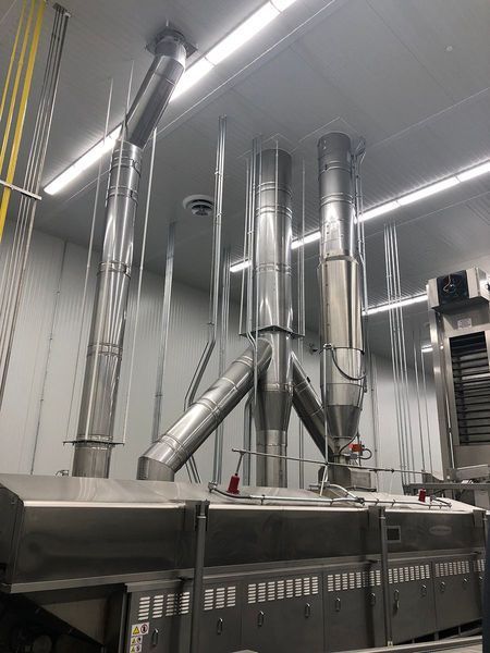 Stainless steel exhaust vents connect to a large industrial food processing machine in a brightly lit facility.