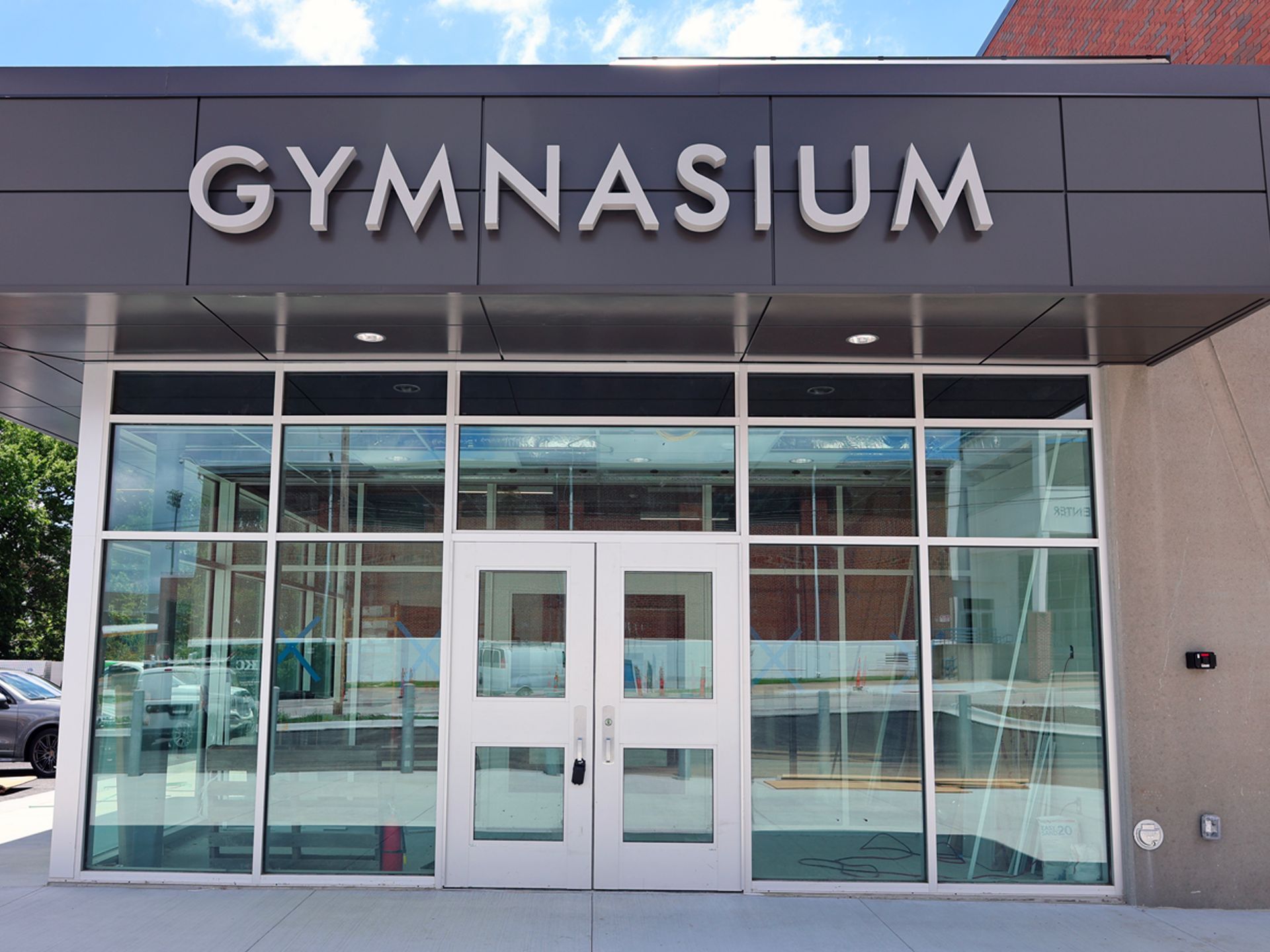 The entrance to a modern gymnasium with large glass windows and the word GYMNASIUM in white letters above the double doors.