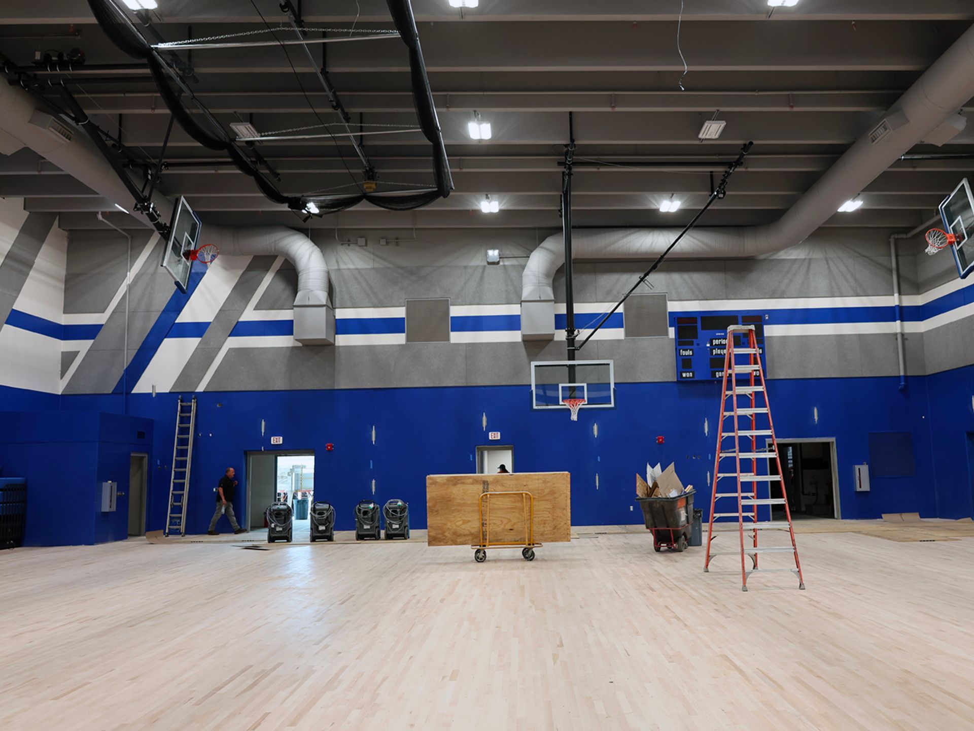 Indoor basketball court under construction with ladders, a wooden platform on wheels, and blue and gray wall accents.