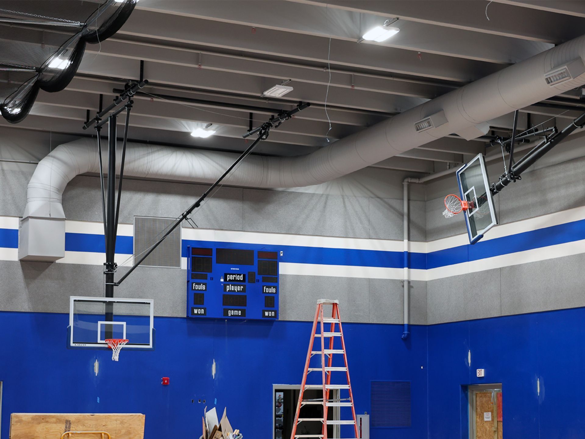 A basketball court interior featuring two hoops, a scoreboard, and an orange ladder against a blue and gray wall.
