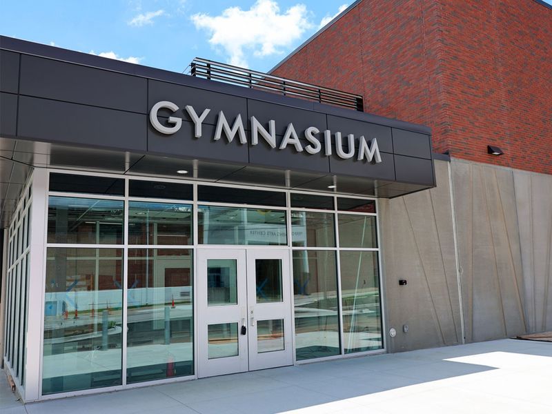Exterior entrance of a modern school gymnasium with glass doors and walls, under a large sign reading 