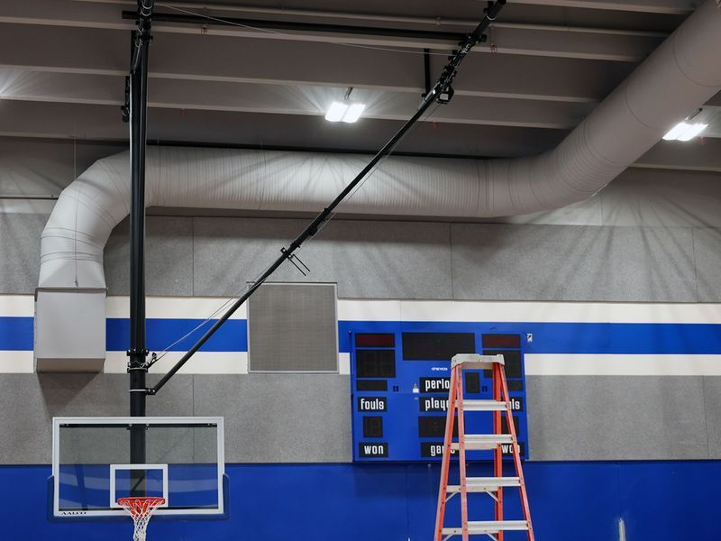 A basketball hoop and scoreboard in a gym, with a ladder standing in front of the wall.