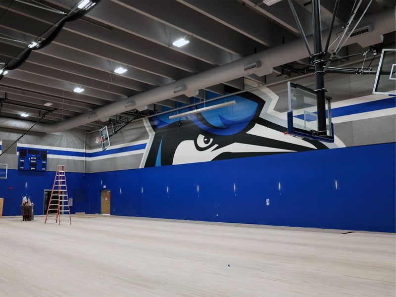 Indoor basketball court with light-colored flooring, blue wall panels, and a large blue jay mascot mural on the back wall.