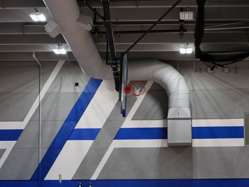 A basketball hoop attached to a wall with geometric gray, white, and blue patterns, located under large HVAC air ducts.